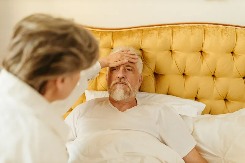 A carer resting a hand on an older man's forehead as he lies in his own bed