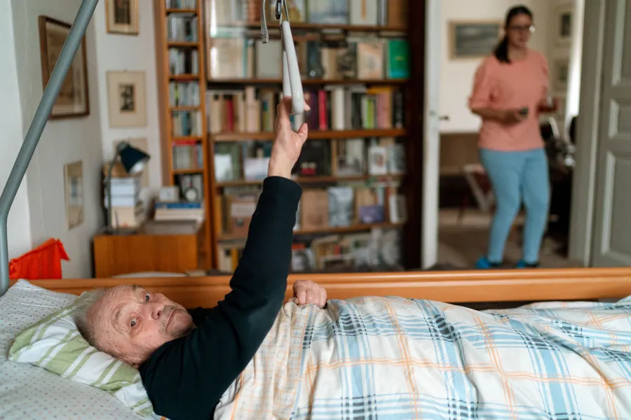 An older man resting in a hospital bed set up in his own book-lined living room